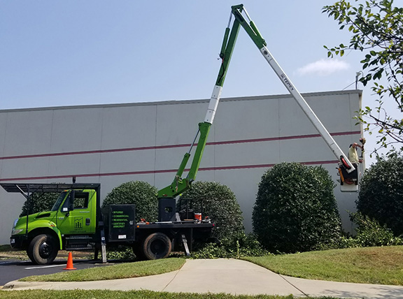 TimberTech bucket truck with an extended boom lift trimming hedges beside a commercial building